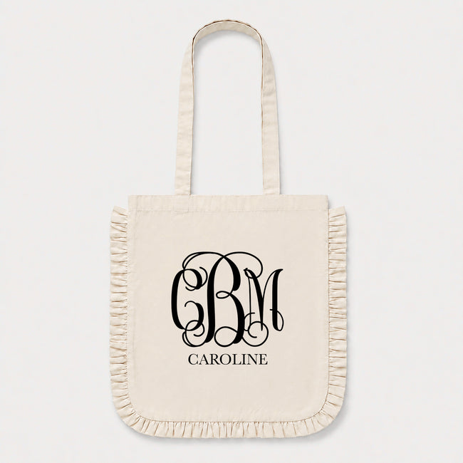 An off-white canvas tote bag with fringed edges and long handles, featuring a black monogram 'CBM' and the name 'CAROLINE' printed on the front, set against a light gray background.