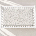 A white wooden baby crib with a fitted sheet printed with an all-over lowercase alphabet pattern in gray, shown from a top-down angle on a textured cream rug.