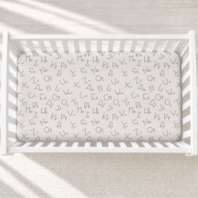 A white wooden baby crib with a fitted sheet printed with an all-over lowercase alphabet pattern in gray, shown from a top-down angle on a textured cream rug.