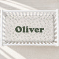 A white wooden baby crib with a fitted sheet printed with small lowercase alphabet letters and the name 'Oliver' in large dark green text across the center, shown from above on a light textured floor.