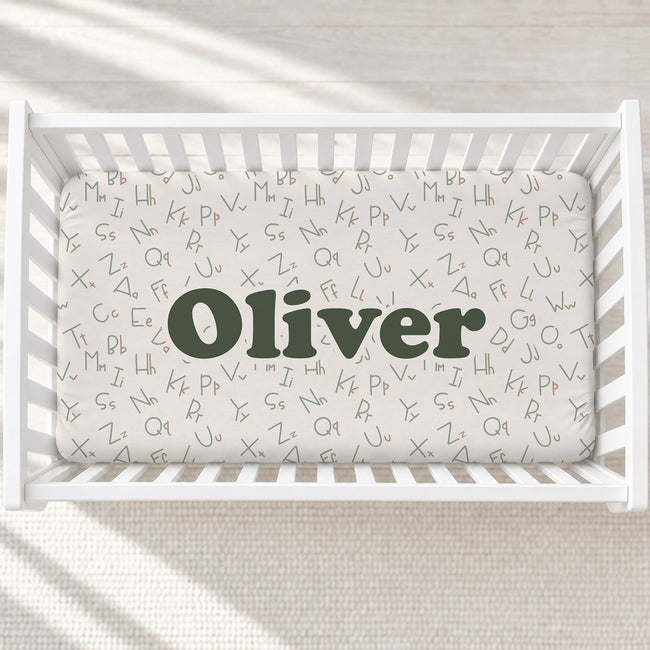 A white wooden baby crib with a fitted sheet printed with small lowercase alphabet letters and the name 'Oliver' in large dark green text across the center, shown from above on a light textured floor.