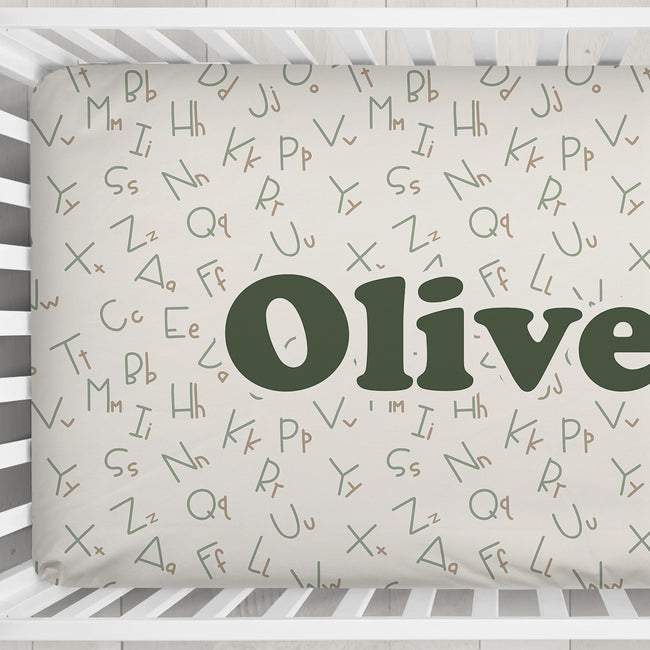 Custom baby crib bedding featuring an alphabet pattern with the name 'Olive' in large dark green letters on a cream background, shown inside a white wooden crib.