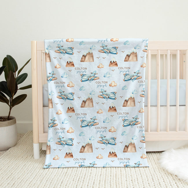 A personalized baby blanket with a repeating pattern of blue airplanes, brown mountains, and the name 'COLTON Joseph' printed in brown, draped over a light wood crib in a nursery setting.