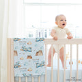 A baby stands in a light wood crib, holding the railing while wearing a cream bodysuit. A matching crib bedding set with a blue sky, airplane, cloud, and mountain pattern is draped over the side of the crib.