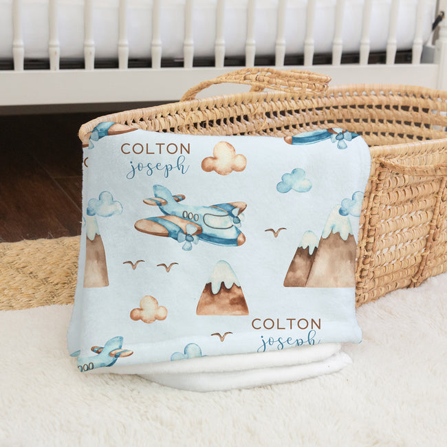 A personalized baby blanket with an airplane and mountain pattern, folded and displayed next to a woven basket on a white rug.