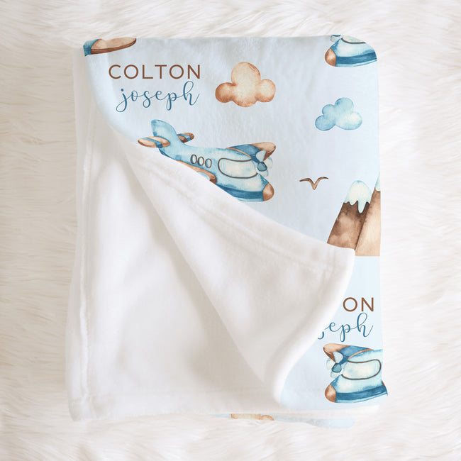 A personalized baby blanket with a white reverse side and a light blue printed front featuring watercolor-style airplanes, clouds, mountains, and the names 'COLTON joseph' in brown and blue lettering, laid flat on a white faux fur surface.