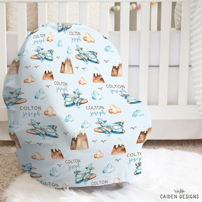 A custom baby car seat cover featuring a light blue background with a watercolor pattern of airplanes, mountains, clouds, and the names 'COLTON joseph' in brown, styled in a nursery setting on a white rug in front of a crib.