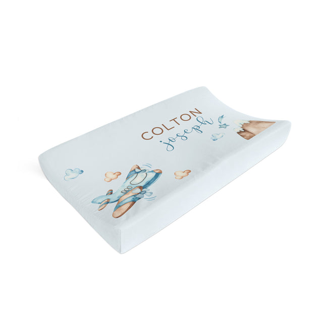 A custom contoured baby changing pad cover, personalized with the name 'COLTON joseph' in brown and blue script, decorated with a watercolor illustration of a blue and brown airplane, on a clean white background.