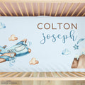 Personalized baby bedding set in a wooden crib, featuring a white sheet with watercolor illustrations of a blue airplane, clouds, stars, and mountains, along with the custom text 'COLTON joseph' in brown and blue lettering.