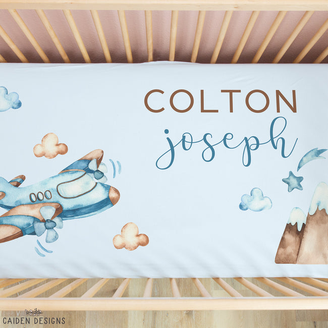 Personalized baby bedding set in a wooden crib, featuring a white sheet with watercolor illustrations of a blue airplane, clouds, stars, and mountains, along with the custom text 'COLTON joseph' in brown and blue lettering.