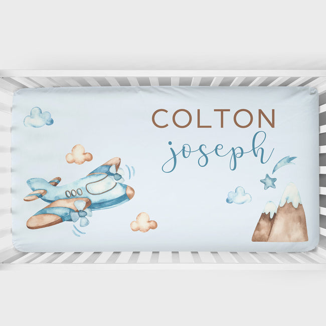 Personalized baby bedding set in a white crib, featuring a light blue background with watercolor illustrations of a blue airplane, clouds, stars, and a mountain. The text 'COLTON' is printed in brown block letters, and 'joseph' is written in blue cursive script.