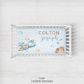 A personalized baby crib sheet with a watercolor design, shown inside a white wooden crib. The sheet features a light blue background with a blue airplane, brown clouds, a brown mountain, and the names 'COLTON joseph' in brown and blue lettering.