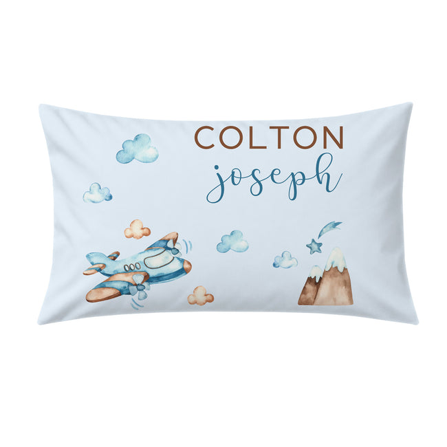A personalized baby pillowcase featuring a light blue watercolor design with an airplane, clouds, a mountain, and the names 'COLTON' and 'joseph' printed on the front.