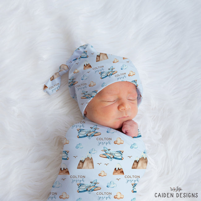 A sleeping newborn baby swaddled and wrapped in a light blue fabric with a printed pattern of mountains, clouds, birds, and the name 'COLTON joseph', wearing a matching tie-on hat. The baby is lying on a white faux fur blanket.