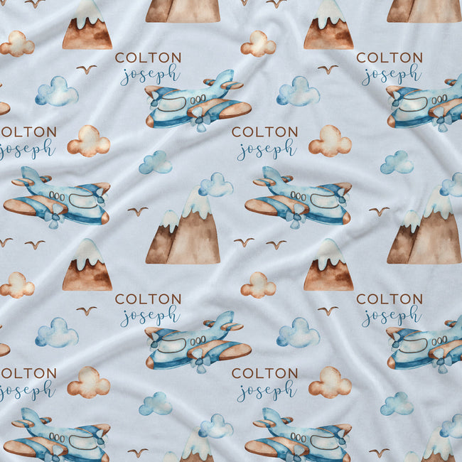 Personalized baby blanket with a repeating pattern of blue watercolor airplanes, brown mountains, beige clouds, and the name 'COLTON joseph' printed in brown and blue text on a white background.