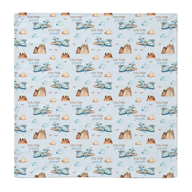 A square swaddle blanket featuring a repeating watercolor pattern of blue airplanes, brown mountains, and beige clouds on a light blue background, with the name 'COLTON joseph' printed repeatedly.