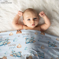 A baby lying on a white faux fur blanket, wrapped in a personalized light blue muslin blanket printed with blue airplanes, brown clouds, and the name 'COLTON joseph'. The brand 'CAIDEN DESIGNS' is visible in the top left corner.