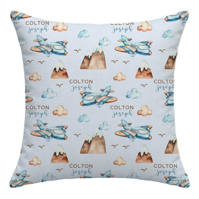 A square throw pillow with a light blue background featuring a repeating watercolor pattern of blue airplanes, brown mountains, beige clouds, and the custom text 'COLTON joseph'.