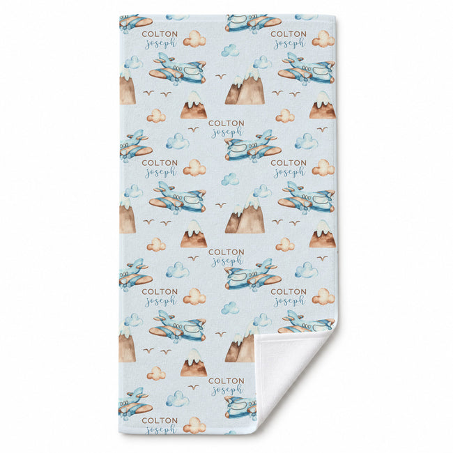 A personalized baby blanket with a light blue background, featuring a repeating pattern of blue airplanes, brown mountains, beige clouds, and the names 'COLTON joseph' printed in brown. The bottom corner is folded over to show the white reverse side.