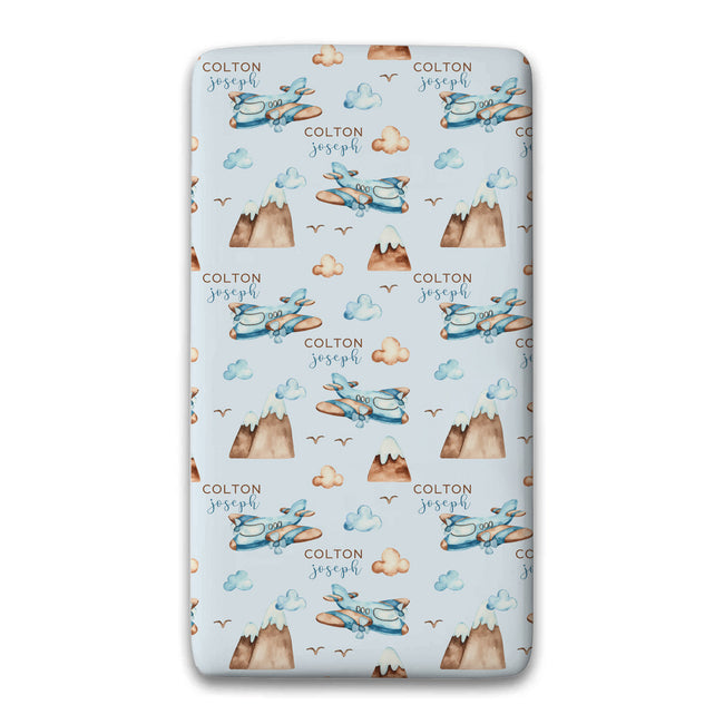 A twin fitted sheet featuring a repeating pattern of blue airplanes, brown mountains, and clouds on a light blue background. The pattern is personalized with the names 'COLTON joseph' printed repeatedly.