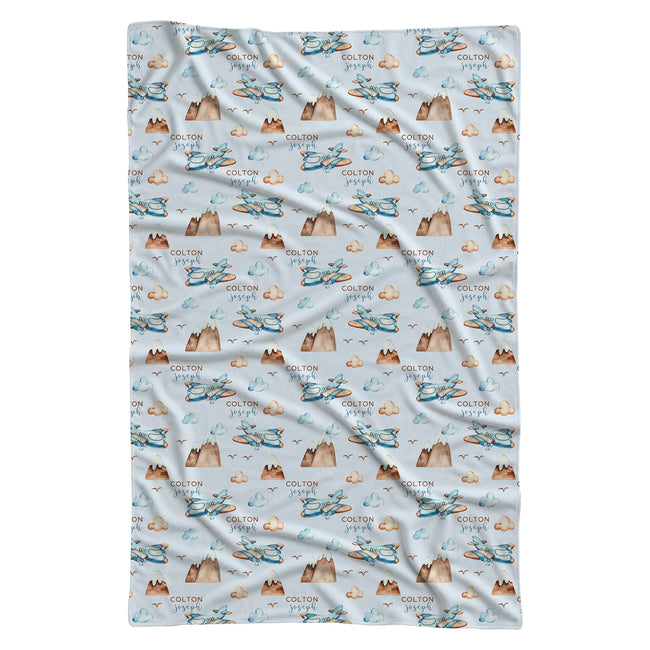 A rectangular swaddle blanket with a repeating pattern of blue and brown mountains, clouds, birds, and the text 'COLTON Joseph' on a light blue background.