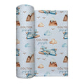 A rolled baby swaddle blanket with a light blue background, featuring a repeating pattern of watercolor-style blue airplanes, brown mountains, beige clouds, and the text 'COLTON joseph' in brown and blue. The product is shown against a plain white background.