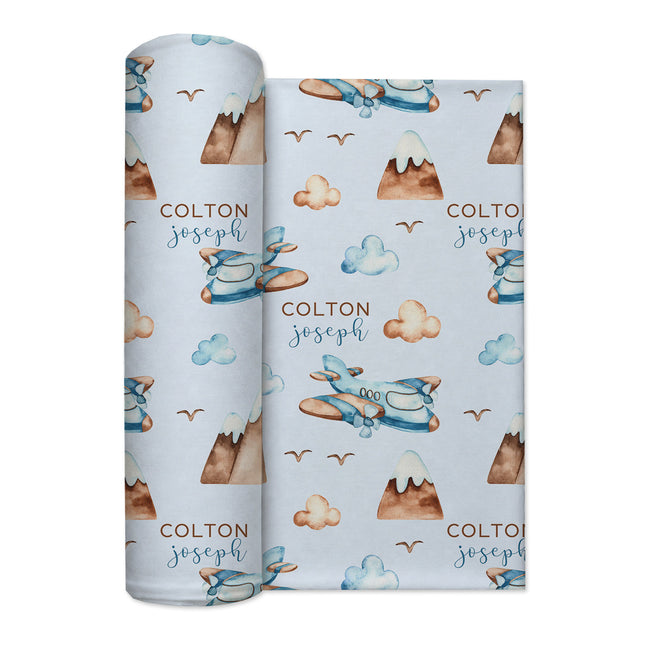 A rolled baby swaddle blanket with a light blue background, featuring a repeating pattern of watercolor-style blue airplanes, brown mountains, beige clouds, and the text 'COLTON joseph' in brown and blue. The product is shown against a plain white background.