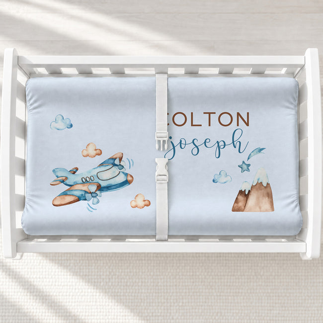A white changing table with a personalized changing pad cover featuring a light blue background with a watercolor airplane, clouds, mountain, and custom names 'COLTON Joseph' in brown and blue lettering.