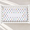 A white crib mattress fitted sheet with a colorful alphabet pattern, shown inside a white wooden crib on a light textured floor.