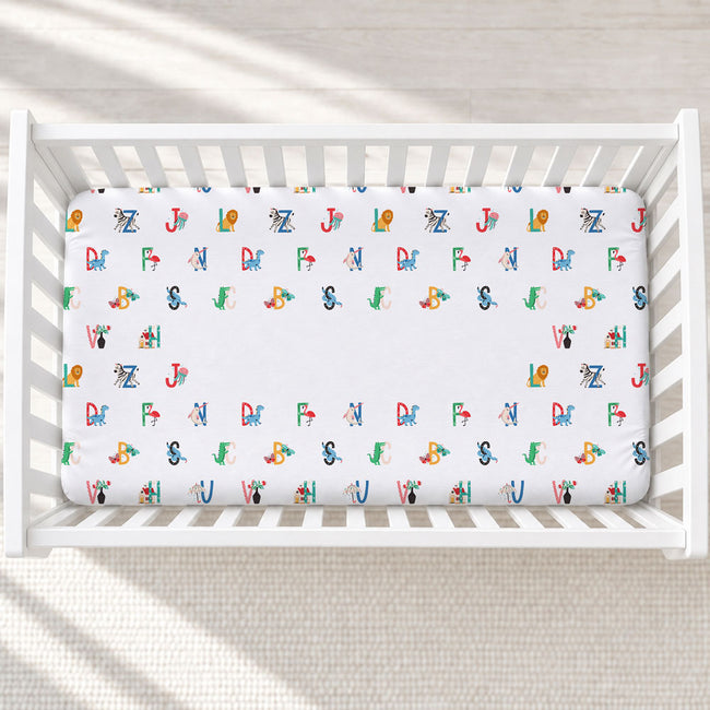A white crib mattress fitted sheet with a colorful alphabet pattern, shown inside a white wooden crib on a light textured floor.