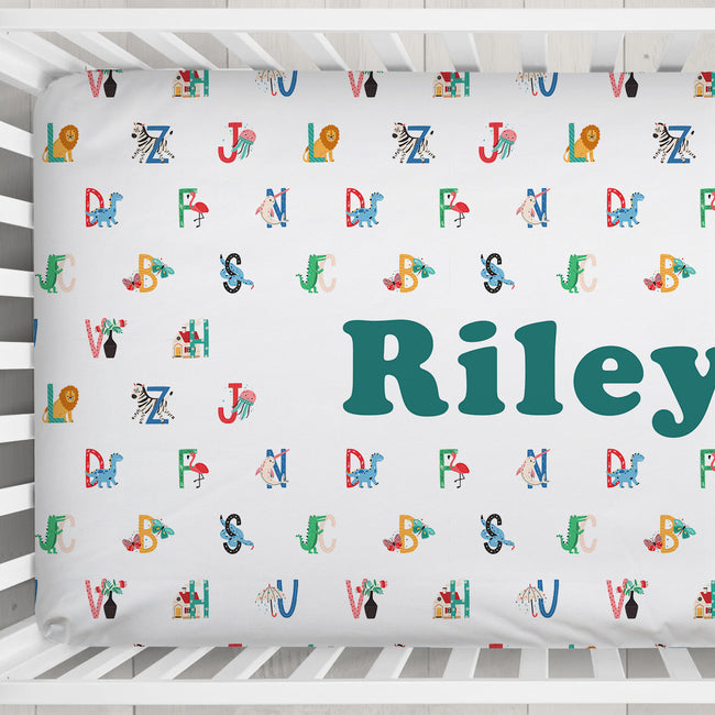 Personalized baby crib bedding set featuring a white fitted sheet and matching pillowcase with colorful alphabet letters, animal illustrations, and the name 'Riley' in large teal text.