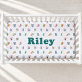 A white crib mattress fitted sheet with a colorful alphabet pattern and the name 'Riley' in large teal letters, shown inside a white crib on a light textured floor.