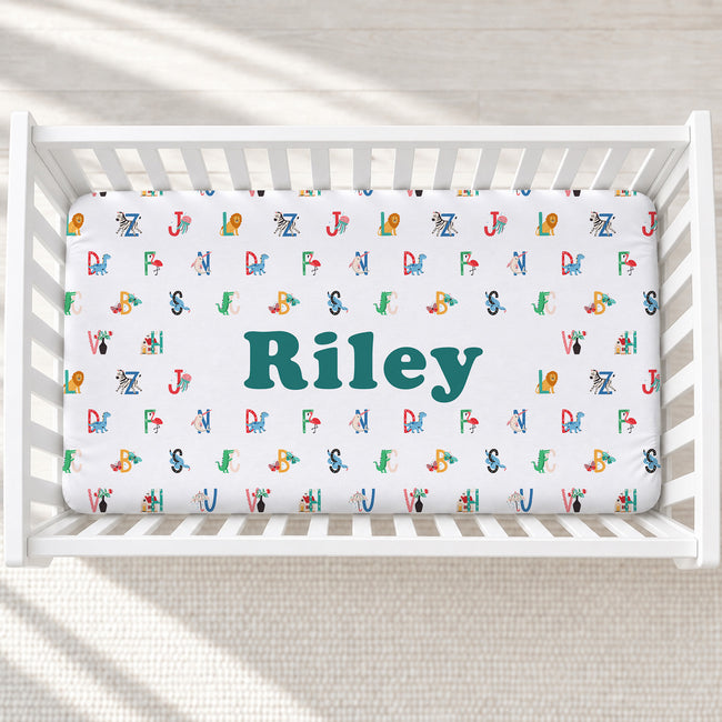 A white crib mattress fitted sheet with a colorful alphabet pattern and the name 'Riley' in large teal letters, shown inside a white crib on a light textured floor.