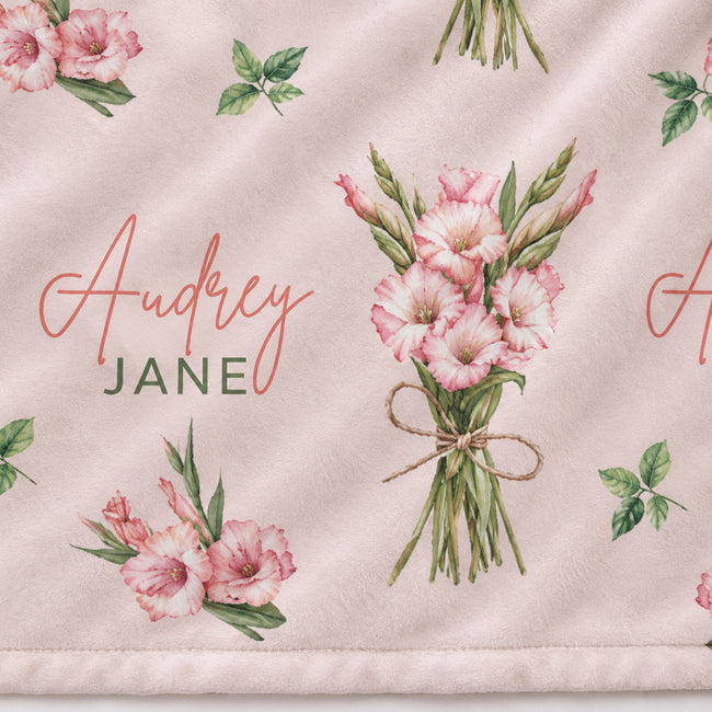A close-up of a light blush pink fabric with a repeating floral print of pink gladiolus flowers and green leaves, plus the personalized name 'Audrey JANE' in coral script and green block letters.