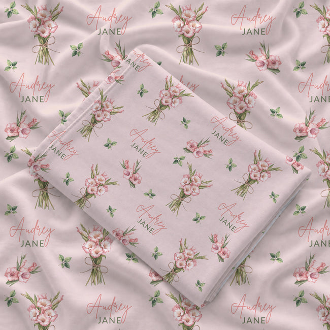 Blush pink swaddle blanket with pink gladiolus bouquets, scattered green leaves, and the name Audrey Jane repeated throughout.