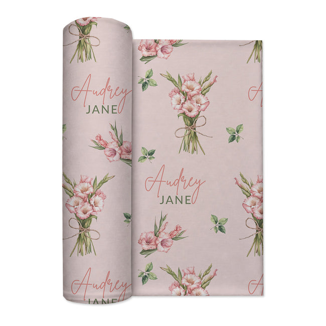 Blush pink swaddle blanket with pink gladiolus bouquets, scattered green leaves, and the name Audrey Jane repeated throughout.