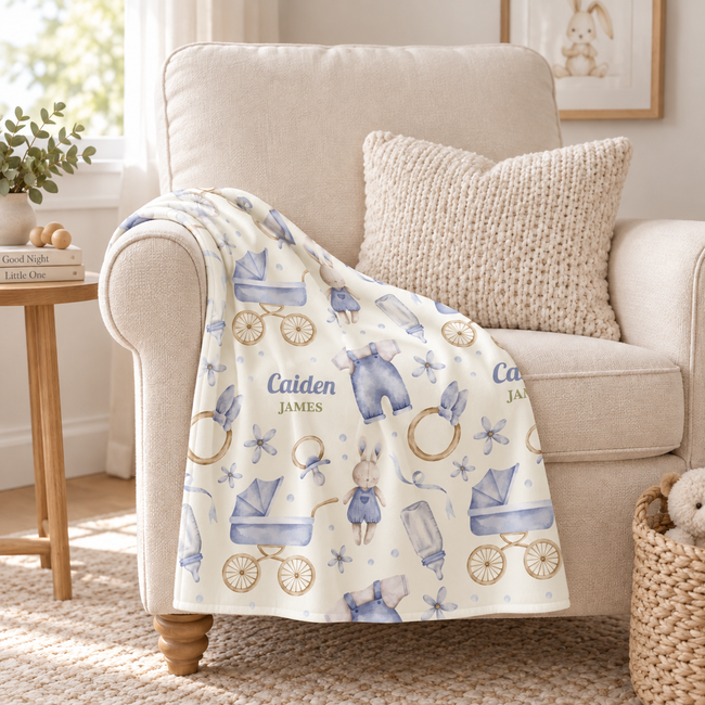A cream-colored throw blanket draped over a beige armchair in a bright, sunlit room. The blanket features a watercolor-style print of baby strollers, buggies, starfish, and small figures in dusty blue, with the names 'Calden' and 'JAMES' printed on it.