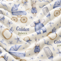 A close-up of a soft cream-colored blanket with a watercolor-style print of baby strollers, bottles, bunnies, and flowers, featuring the personalized name 'Caiden James' in blue and green text.