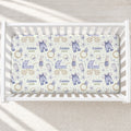 A personalized baby crib mattress sheet with a watercolor-style print of blue baby clothes, strollers, bicycles, rabbits, and gold rings on a cream background, shown inside a white wooden crib.
