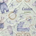 Personalized baby fabric with a watercolor-style print featuring a bunny in blue overalls, a baby stroller, pacifier, bottle, rings, and flowers on a cream background. The text 'Caiden JAMES' is printed in blue and green on the right side.