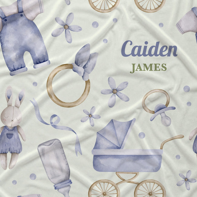 Personalized baby fabric with a watercolor-style print featuring a bunny in blue overalls, a baby stroller, pacifier, bottle, rings, and flowers on a cream background. The text 'Caiden JAMES' is printed in blue and green on the right side.