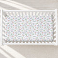 A top-down view of a white wooden crib containing a fitted sheet with a whimsical dinosaur and safari print in soft pastel colors, personalized with the name 'adam' repeated across the fabric.