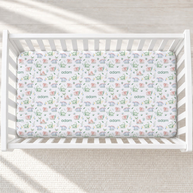 A top-down view of a white wooden crib containing a fitted sheet with a whimsical dinosaur and safari print in soft pastel colors, personalized with the name 'adam' repeated across the fabric.