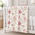 A personalized baby blanket draped over a white crib in a bright nursery. The blanket has a cream background with a pattern of pink ballerina dresses, pink strollers, baby bottles, and the names 'Olivia' and 'Grace' printed in a dusty rose font.