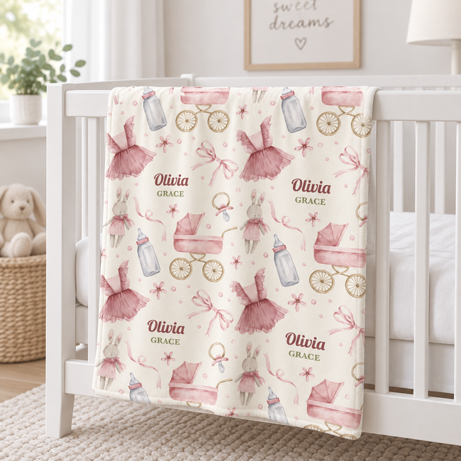 A personalized baby blanket draped over a white crib in a bright nursery. The blanket has a cream background with a pattern of pink ballerina dresses, pink strollers, baby bottles, and the names 'Olivia' and 'Grace' printed in a dusty rose font.
