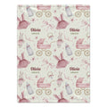 A personalized baby blanket with a repeating pattern of pink prams, bottles, bunnies, and bows on a cream background, featuring the custom text 'Olivia GRACE' in the center.