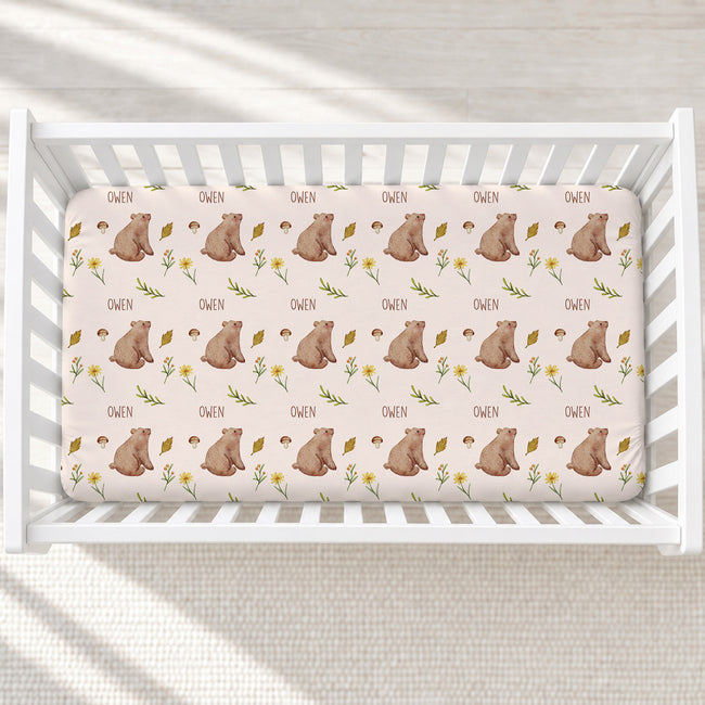 A fitted crib mattress with a repeating pattern of brown teddy bears, yellow flowers, and the name 'OWEN' printed in black, shown inside a white wooden crib.