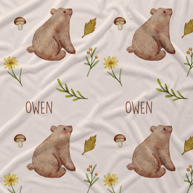 Personalized fabric print featuring a repeating pattern of brown watercolor bears, yellow flowers, mushrooms, and green leaves on a cream background, with the name 'OWEN' printed in brown text.