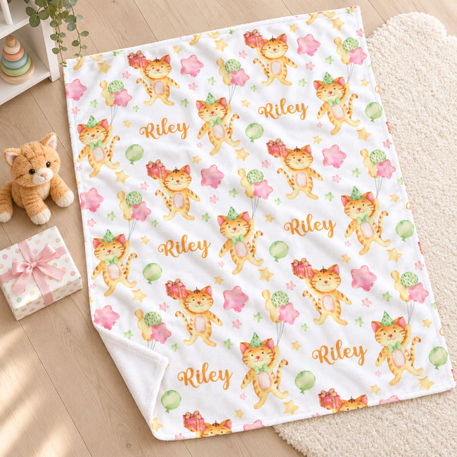 A personalized baby blanket with a white background, printed with cartoon orange tabby cats, pink stars, green balloons, and the name 'Riley' in gold script. The blanket is styled on a light wood floor next to a plush toy, a gift box, and a soft cream rug.
