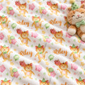 A close-up of a white fabric with a repeating print of orange cartoon cats wearing party hats, pink stars, green balloons, and the name Riley in orange script. A plush teddy bear with a green bow tie is partially visible on the right side.